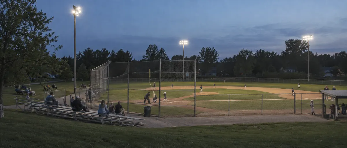A youth baseball field at dusk with the lights on and a few families in the stands